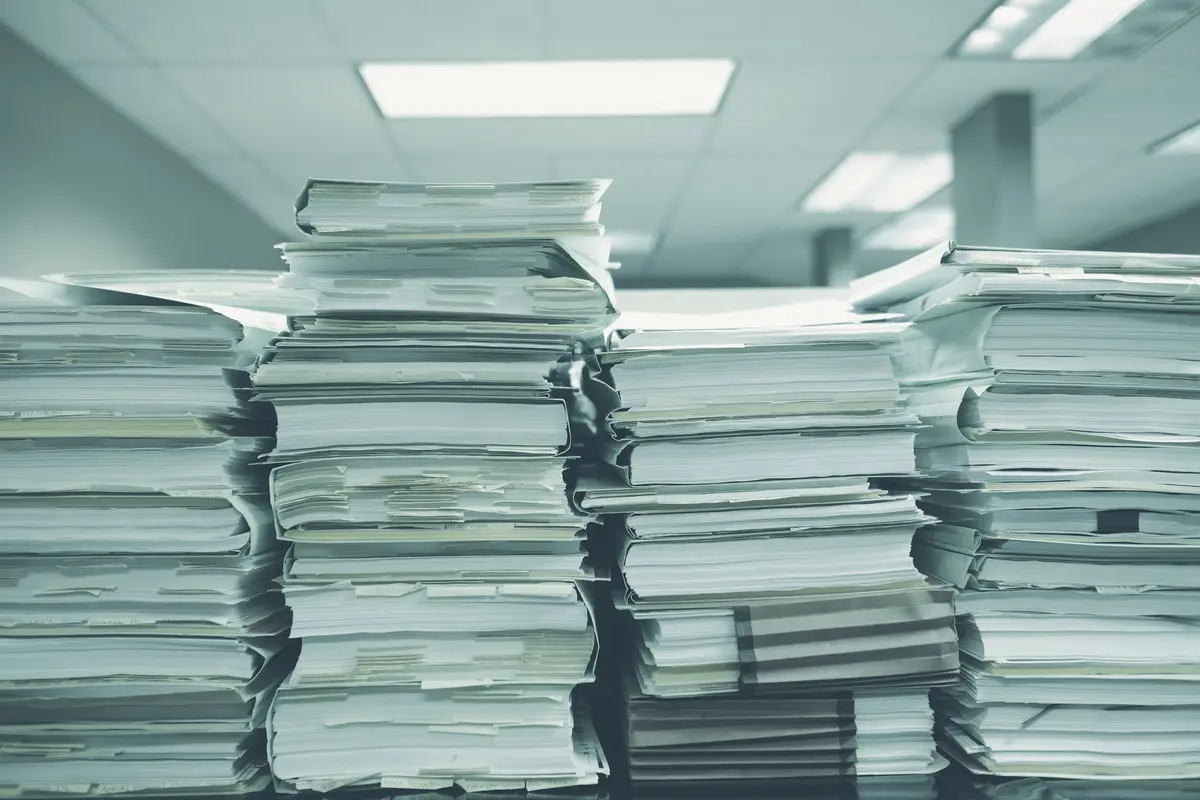 Tall stacks of paper files and folders piled on a table in an office, a picture of records that exist but are hard to use